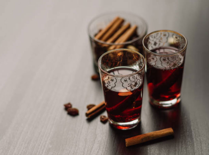 Three clear glasses of glogg sit on a dark table with a bowl of cinnamon sticks. It looks like a warm hygge welcome.