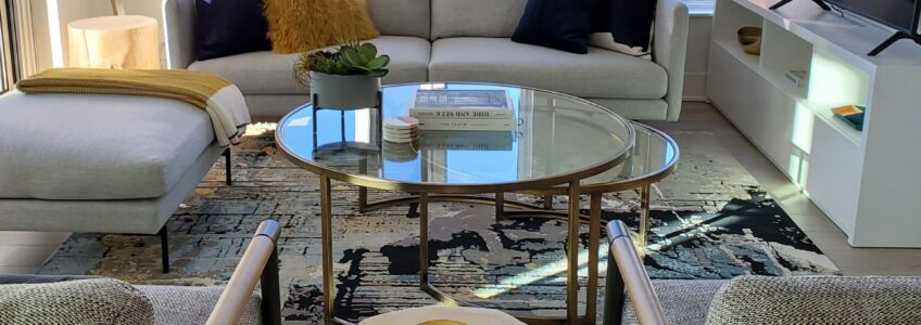A Scandinavian and modern style condo living room. Scandinavian chairs sit across from a low profile simple sofa and ottoman. Nesting glass tables with gold accents. Interesting futuristic floor lamps.