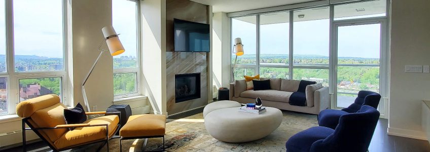 A floor to ceiling light onyx fireplace is the focal point of this room. A large colourful rug makes this space. The bright yellow lounge chair, light neutral sofa, two neutral river stone shaped coffee tables brighten this space while the rich royal blue velvet accent chairs play off the other dark features of the space.
