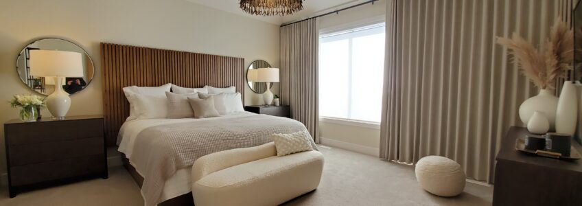 A cozy neutral bed is loaded with textural blankets and pillows. The headboard is a rich wood coloured paneling. White large scale lamps sit on dark night stands and a circular mirror is hung behind to amplify their soft light.