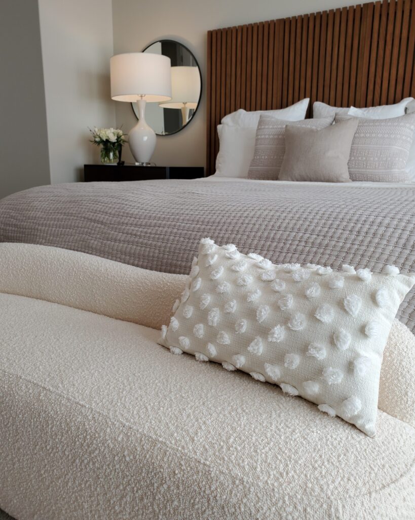 This ensuite has the balance of luxurious high quality features and cozy hygge elements. This angle of photo shows a soft boucle bench at the foot of a king sized bed loaded with plush pillows. All in a neutral palette with wood accents.
