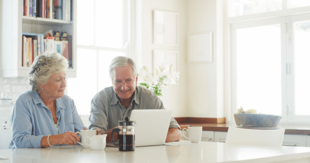 Aging in place - a man and a woman with silver hair sit at their kitchen table enjoying a cup of coffee and looking at their laptop. The kitchen is white and bright.
