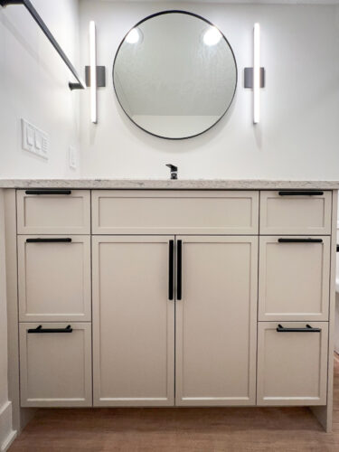 Beige bathroom vanity with circular mirror flanked by two stunning line lighting fixtures.