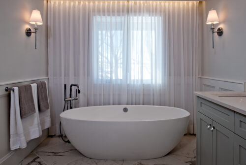White and grey bathroom with standalone oval bathtub with floor mounted faucet. Behind it sheer curtains let in light from a large window, but still give privacy.