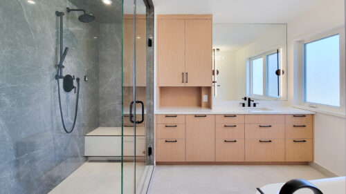 Ensuite bathroom. To the left a grey tiled steam shower complete with a lighter tiled bench. To the right, a warm wood vanity allows for makeup to be done to the left and washing to be done to the right. Line style sconces are mounted to the mirror. Black hardware adorns this look.