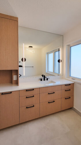 A warm wood vanity allows for makeup to be done to the left and washing to be done to the right. Line style sconces are mounted to the mirror. Black hardware adorns this ensuite bathroom vanity.