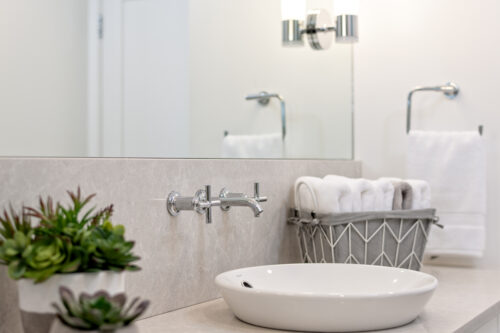 Bathroom vanity with wall-mounted faucets on a raised basin with a grey basket holding clean cloths.