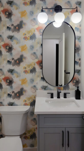 Powder room bathroom with racetrack mirror, blue vanity, black accents and colourful floral wallpaper.