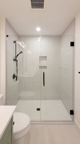 After photo of midcentury renovated bathroom shower. Vertical light grey tiles adorn this oversized shower with glass doors and black accessories.
