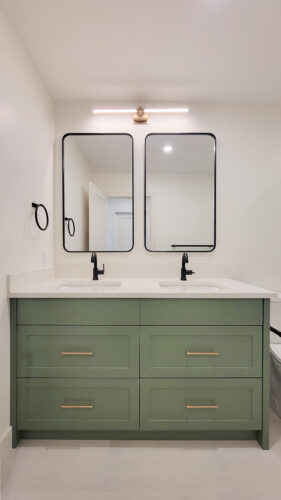 After photo of midcentury renovated bathroom vanity. Green vanity cabinets with gold hardware and double sinks with black accessories. Two separate rounded rectangular mirrors and a lighting fixture that nods to the midcentury origina of this home.