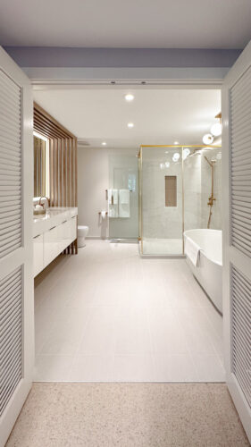 After photos of primary bathroom ensuite in river house. The entry feels like you are going into a light, bright spa with golden and wood accents. To the left is the white vanity with a decorative panel dividing the toilet from the rest of the space. To the right is a glass encased shower with gold accents and light marble look tiles with a wooden look tiled niche. Also to the right is a standalone white tub.