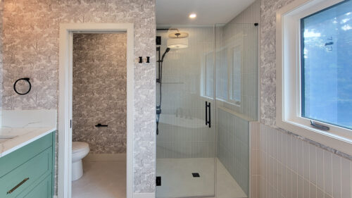 Ensuite bath with jungle wallpaper on all 4 walls. This view is of the toilet room that has a pocket door and the over sized curbless shower with vertical grey subway tiles.