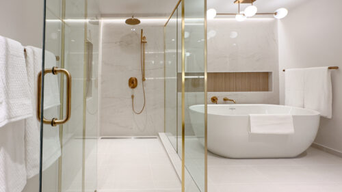 After photo of primary ensuite bathroom in river house. To the left is a glass encased shower with gold accents and light marble look tiles with a wooden look tiled niche. Also to the right is a standalone white tub with a long rectangular wooden-look tiled niche for candles or a wine glass to rest.
