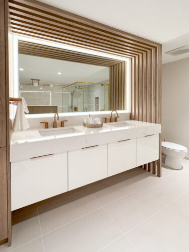 After photo of the primary ensuite bathroom in river house. To the left is a white floating vanity with stone countertops, gold faucets, and a decorative panel going from floor to ceiling, running along the ceiling and over. The vanity light is rectangular and surrounds the entire mirror.
