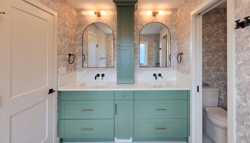 After photo of midcentury and transitional primary ensuite bathroom. Two arched mirrors give a nod to the previous arched mirrors in the sitting room. Each mirror has an wall-mounted light and wall-mounted black faucets over the double sinks. In between the sinks there is a counter to ceiling central medicine cabinet. The wallpaper surrounds all 4 walls and is a playful black and white jungle theme.