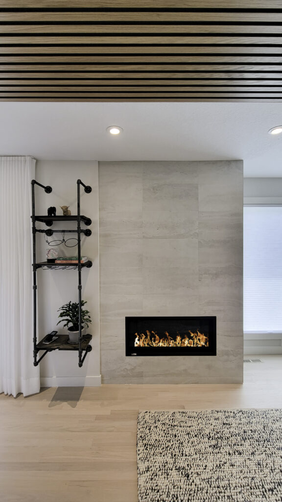 After photo of fireplace covered with a light grey fabricated stone tile. Beside it, a bespoke plumbing pipe shelf holds a client's mementos. The floor is a refreshed and tied in light maple.