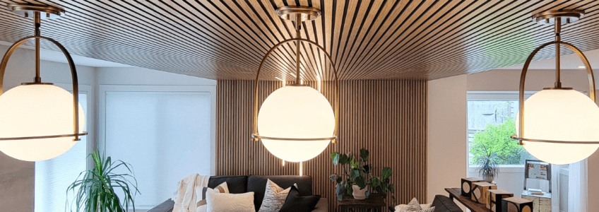 Construction on a renovated home includes striking wooden slats that go from the wall to the ceiling. Three spherical lights hang in the foreground illuminating the living room behind.
