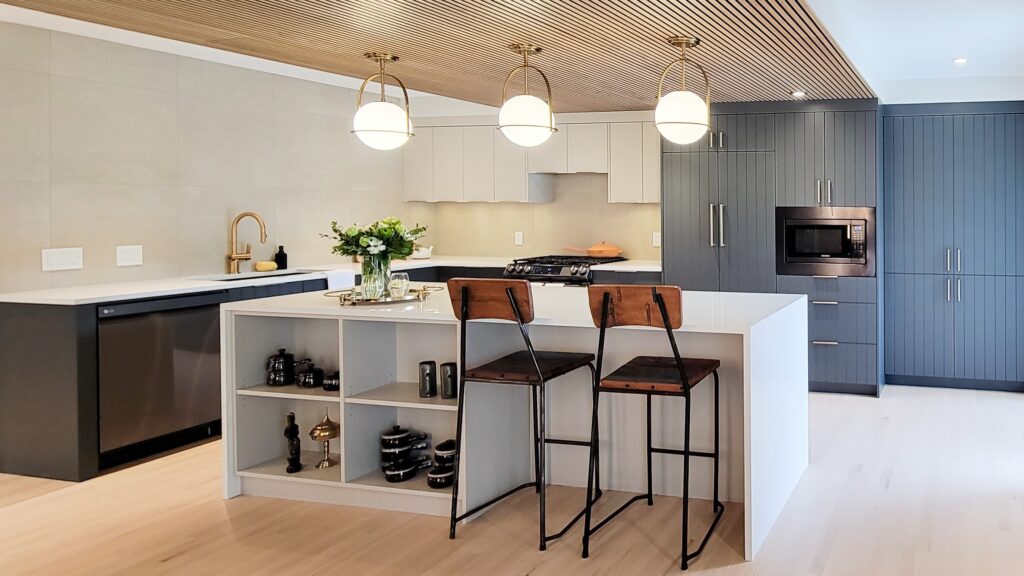 After Construction Kitchen. This kitchen has an architectural detail on the ceiling that includes wooden slats. The wall cabinets are dark blue grey and the island is light white. Two wooden bar stools sit at the island.