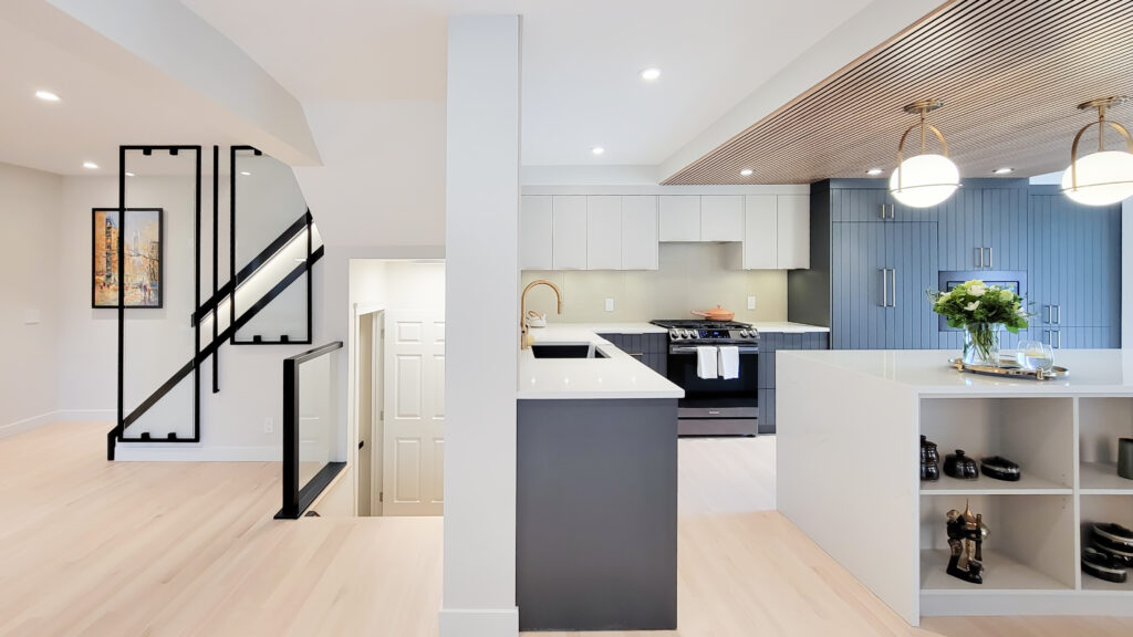 Main floor construction after photo. Stairs have striking glazing with black frames and railings to match. The kitchen has light grey upper cabinets and dark blue grey cabinets and striking globe lights with gold accents.