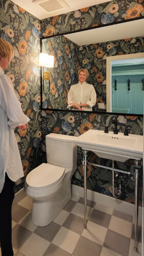 Krista in a powder room that is fully covered in bold floral wallpaper. It almost feels like you are heading down Alison Wonderland's rabbit hole. The pedestal sink has chrome legs. The faucet is matte black. The sconces have linen lamp shades that add to the old world charm.