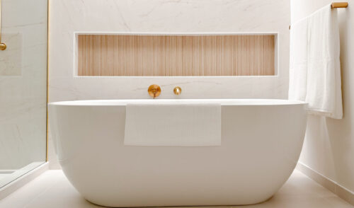 Bathroom ensuite acrylic standalone tub stands in front of an alcove with ribbed wood stained ceramic tile for candles or bath accessories.