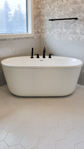 This bathroom ensuite features a gorgeous corner standalone acrylic tub with matte black fixtures. Grey tile in the background up to half the wall height and monochrome forest wallpaper above. A light grey green hexagonal floor tile grounds this look