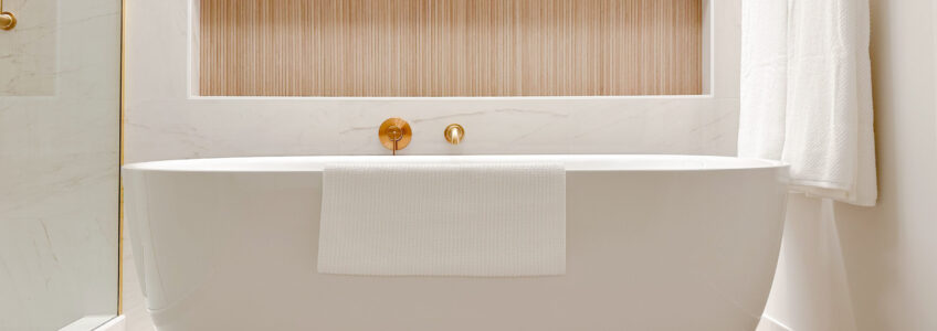 Luxurious standalone bathtub with gold faucet on the marble tiled wall behind. A niche for bath toiletries or candles is inset with wood coloured tiles giving beautiful contrast and a spa-like feeling.