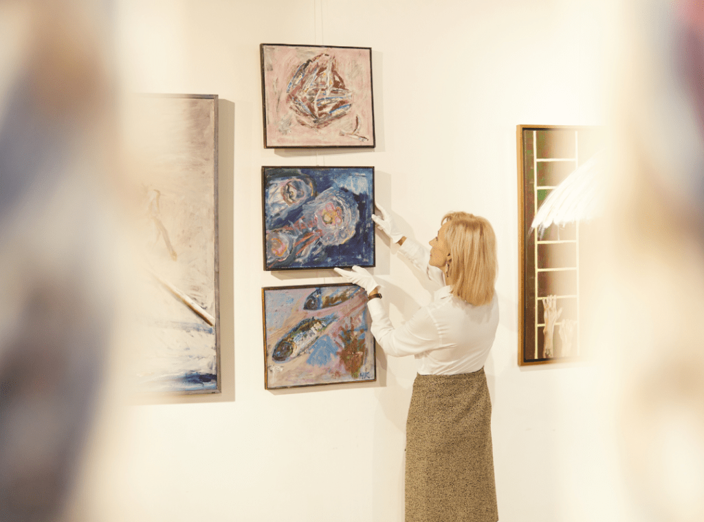 A woman hangs an art piece on a wall with a collection of other works.