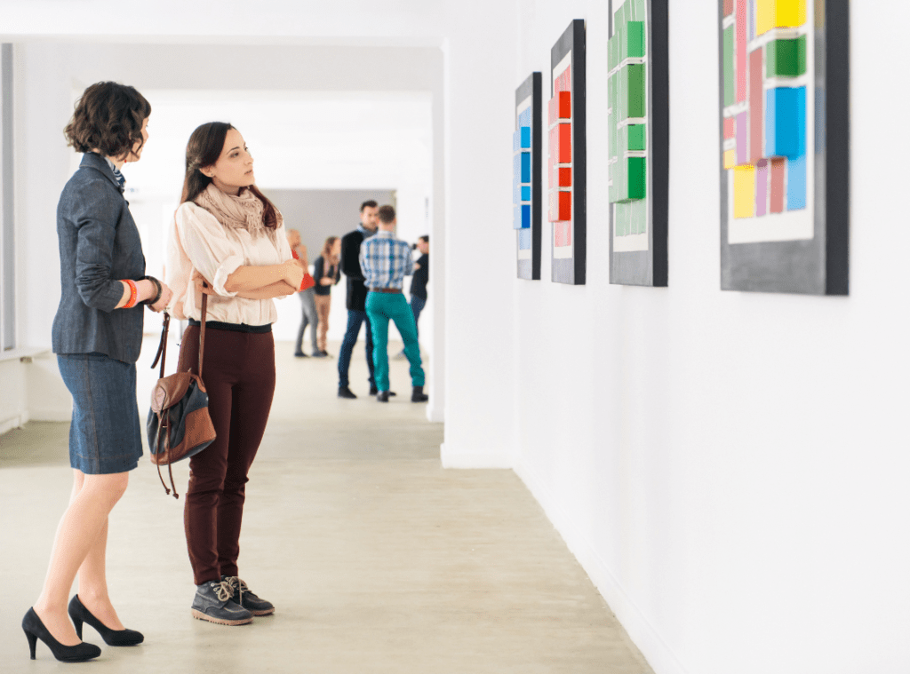 Two women look at art in a gallery. The art is abstract, graphical, and multicoloured with projecting cube and rectangular shapes.
