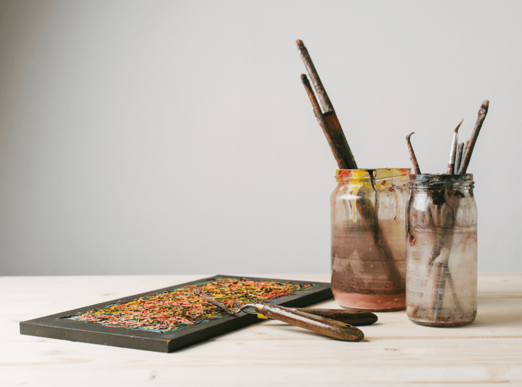 A rectangular paint palette with two palette knives sit on a wooden surface. Two old glass jars filled with dirty paint brushes sit beside it.