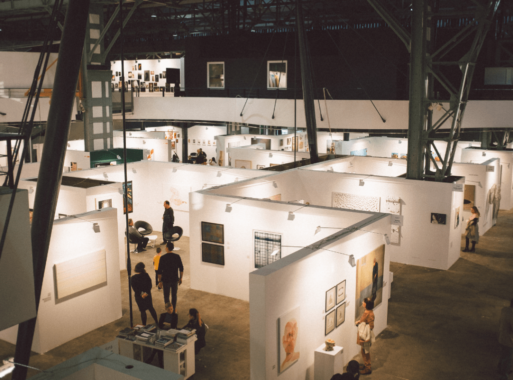 Large warehouse filled with an art exhibit. White makeshift walls separate different artist's work in a maze-like shape.