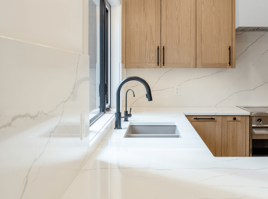 Marble look quartz countertop with subtle but strong veining throughout is carried up the backsplash. A double undermounted sink sits in front of a large window. Black fixtures and instant hot water dispenser adds to the luxury. Light warm wood cabinetry brings it all together.